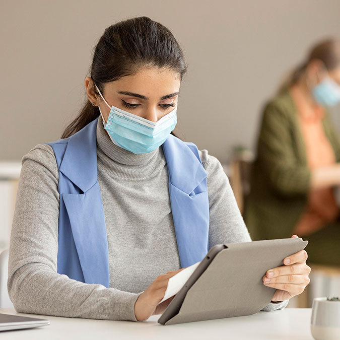 woman with mask sitting at a table holding a tablet